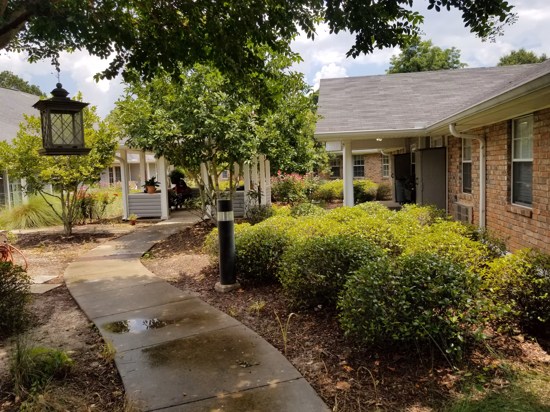 A paved walkway curves through a garden area with green bushes and trees, leading to a covered seating area with benches. The surrounding buildings have brick walls and windows, and the sky is partly cloudy.