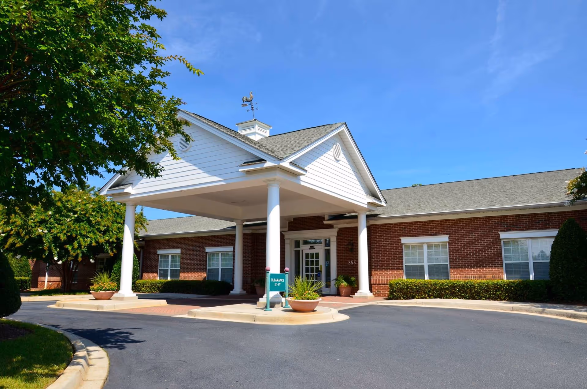 Front entrance of a one-story brick senior living building with a white portico, columns, potted plants, and a circular drive-up.