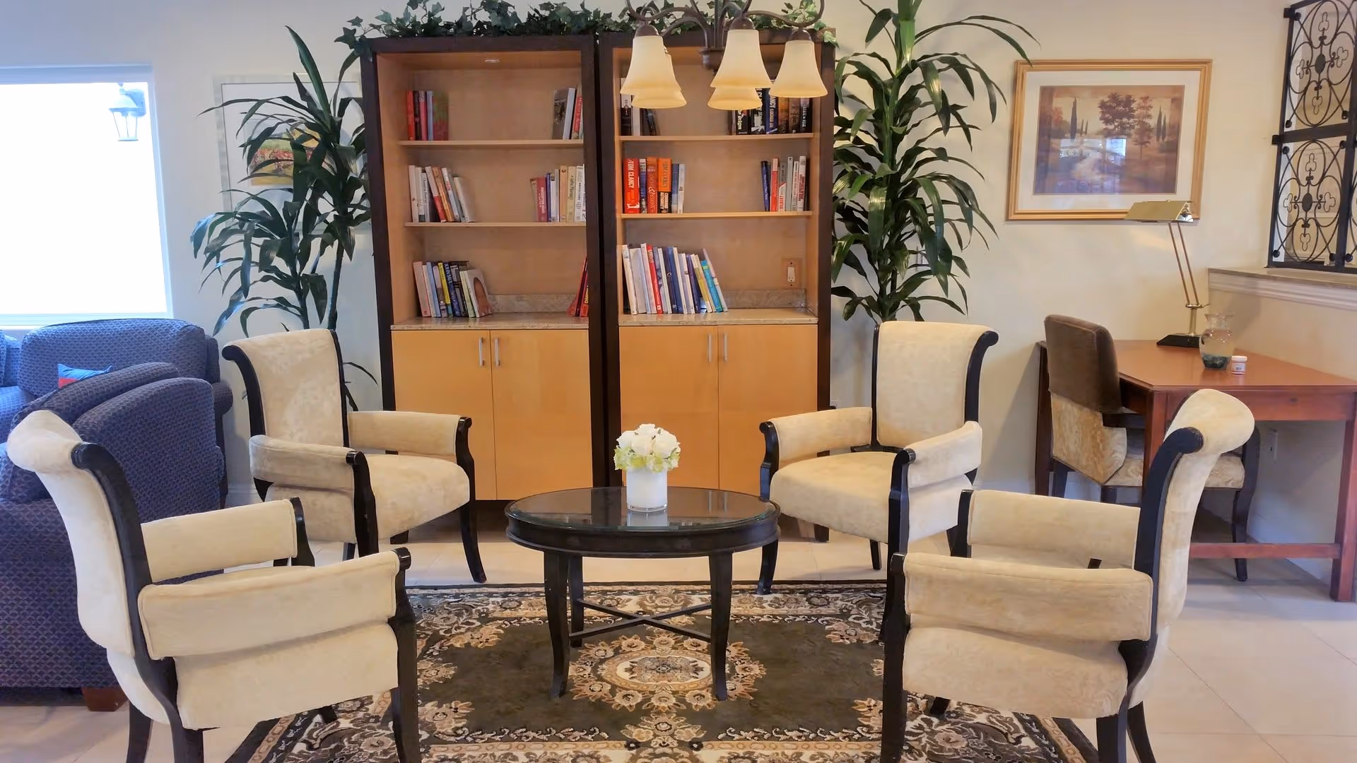 A cozy sitting area in an assisted living facility featuring four beige upholstered armchairs arranged around a round black coffee table with a small flower arrangement. Behind the chairs are two wooden bookshelves filled with books, flanked by tall green plants. To the right, there is a wooden desk with a chair and a lamp, and a framed artwork hangs on the wall.