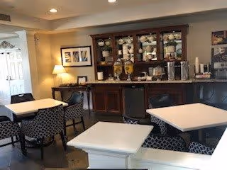 Interior view of a dining area with several square white tables and patterned chairs. A wooden cabinet with glass doors displays dishes and glassware, and there are beverage dispensers on the counter below. The room is softly lit with ceiling lights and a table lamp, and framed artwork is hung on the walls.