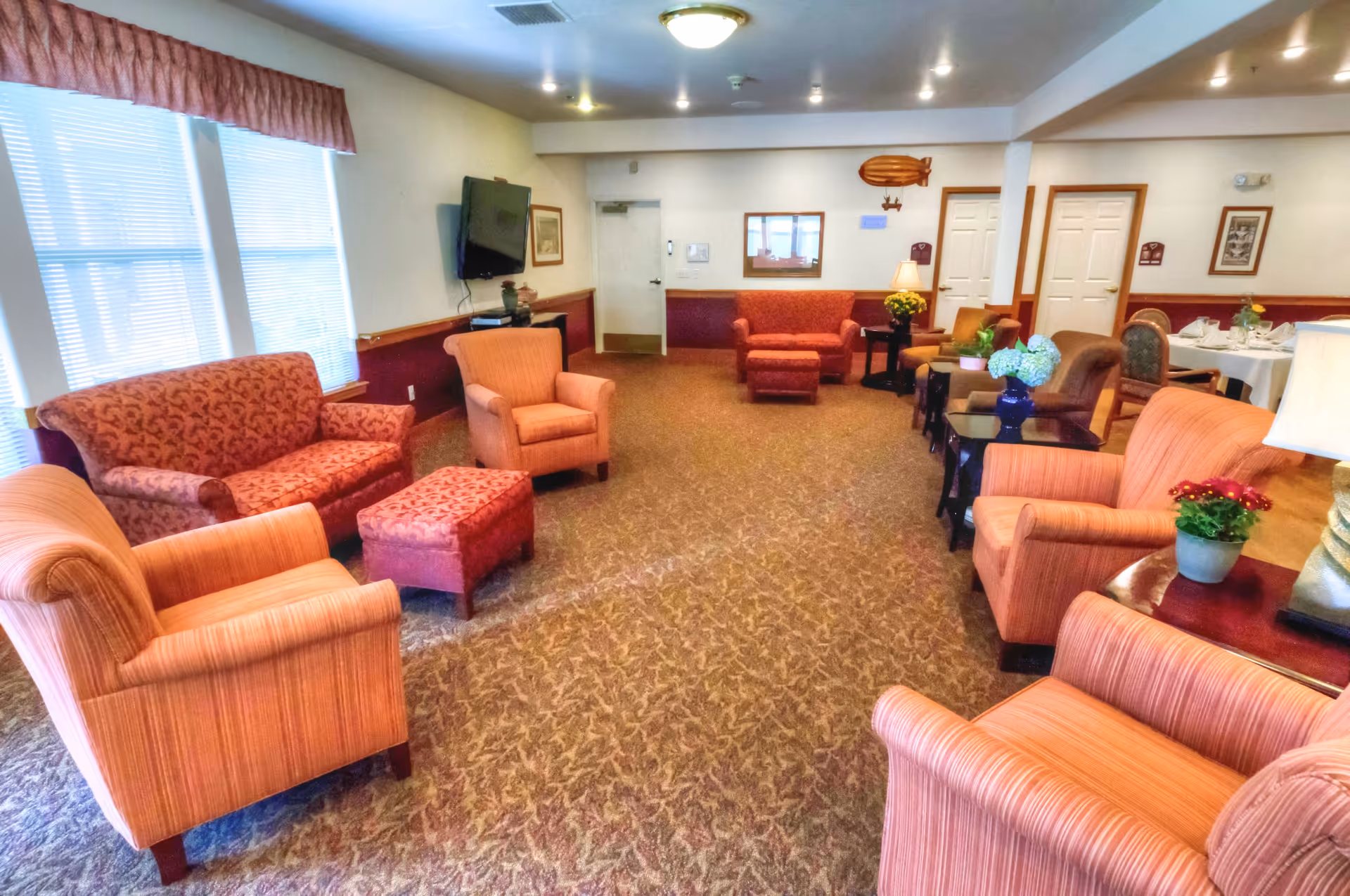 A cozy living room area in a senior living facility with multiple upholstered armchairs and sofas in warm tones arranged around small tables with decorative plants and lamps. The room has carpeted flooring, large windows with blinds and valances, a wall-mounted TV, and a dining area with set tables visible in the background.