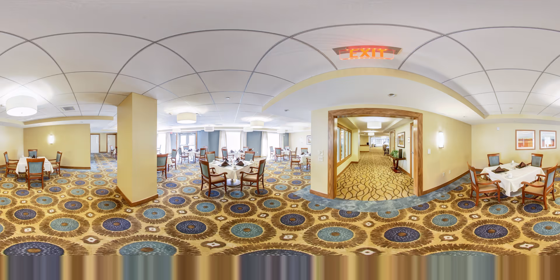 A spacious dining room with multiple round tables covered with white tablecloths and set with napkins and tableware. The room has patterned carpet with circular designs in blue, brown, and beige tones. There are wooden chairs with cushions around the tables. The walls are painted light yellow, and the ceiling has recessed lighting and modern light fixtures. A doorway leads to a hallway with similar carpet and wall decor. An illuminated exit sign is visible above the doorway.