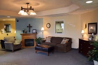 A cozy living room area in a nursing center featuring a brown sofa with patterned pillows, a matching ottoman, a gray armchair, a wooden fireplace with decorative items, a wall clock, table lamps, framed artwork, and indoor plants. The walls are painted in light beige and blue tones, and the room is softly lit by ceiling and table lights.