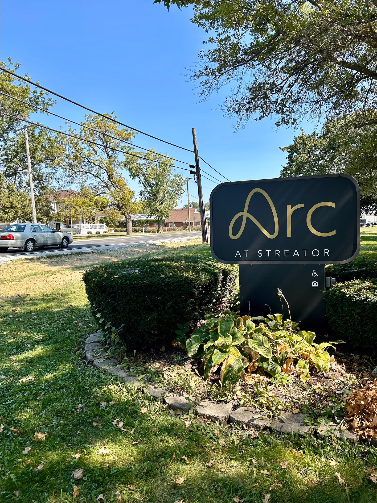A landscaped lawn and shrubs in front of a roadside sign reading "Arc at Streator" with trees and houses beyond.