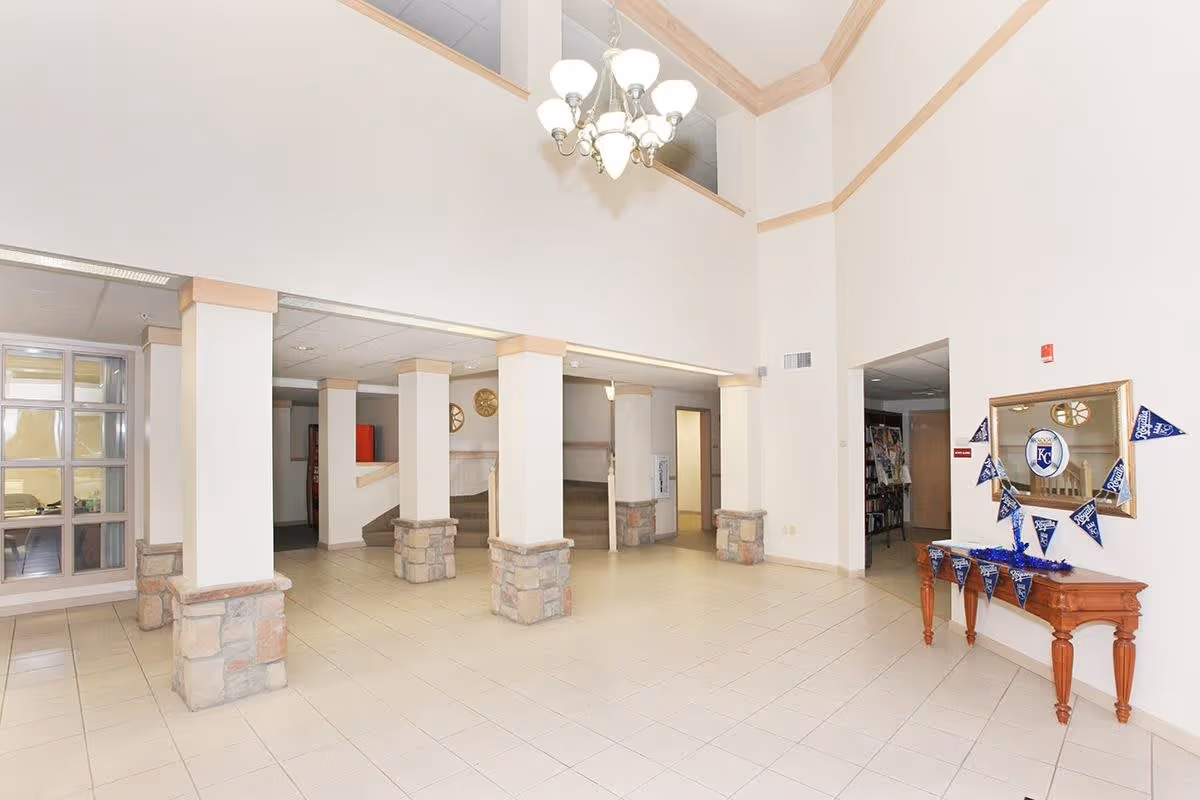A spacious interior lobby area with tiled floors, white walls, and several square columns with stone bases. There is a wooden table on the right side decorated with blue pennants and a mirror above it. A chandelier hangs from the high ceiling, and there are doorways and windows visible in the background.