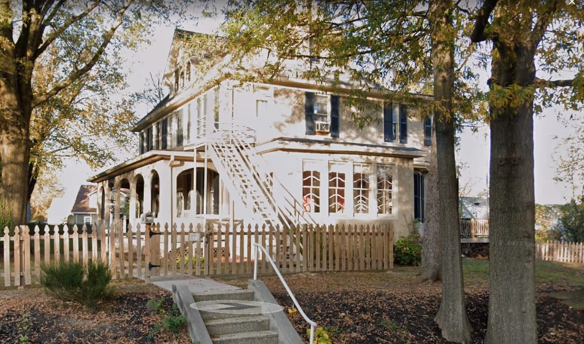 Exterior view of a two-story residential building with a white picket fence, surrounded by large trees and a pathway leading to the entrance. The building has multiple windows and an external staircase on the side.