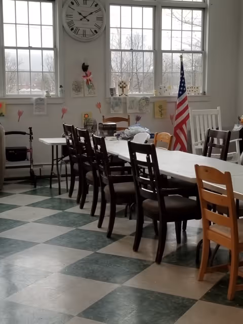 A bright room with large windows showing a view of trees outside. Inside, there is a long table with multiple chairs arranged around it. The floor has a green and white checkered pattern. On the wall above the table, there is a large clock and various decorations including drawings and cutouts. An American flag stands in the corner near a white rocking chair.