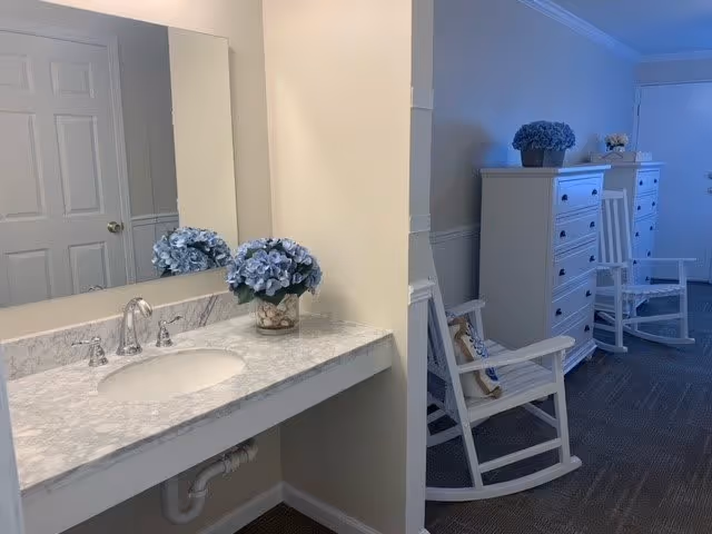 A clean and bright interior space featuring a marble countertop with a sink and silver faucet on the left side, decorated with a vase of blue flowers. To the right, there is a hallway with white rocking chairs and white dressers, each adorned with blue flower arrangements. The walls are painted light beige, and the floor is carpeted in a neutral tone.