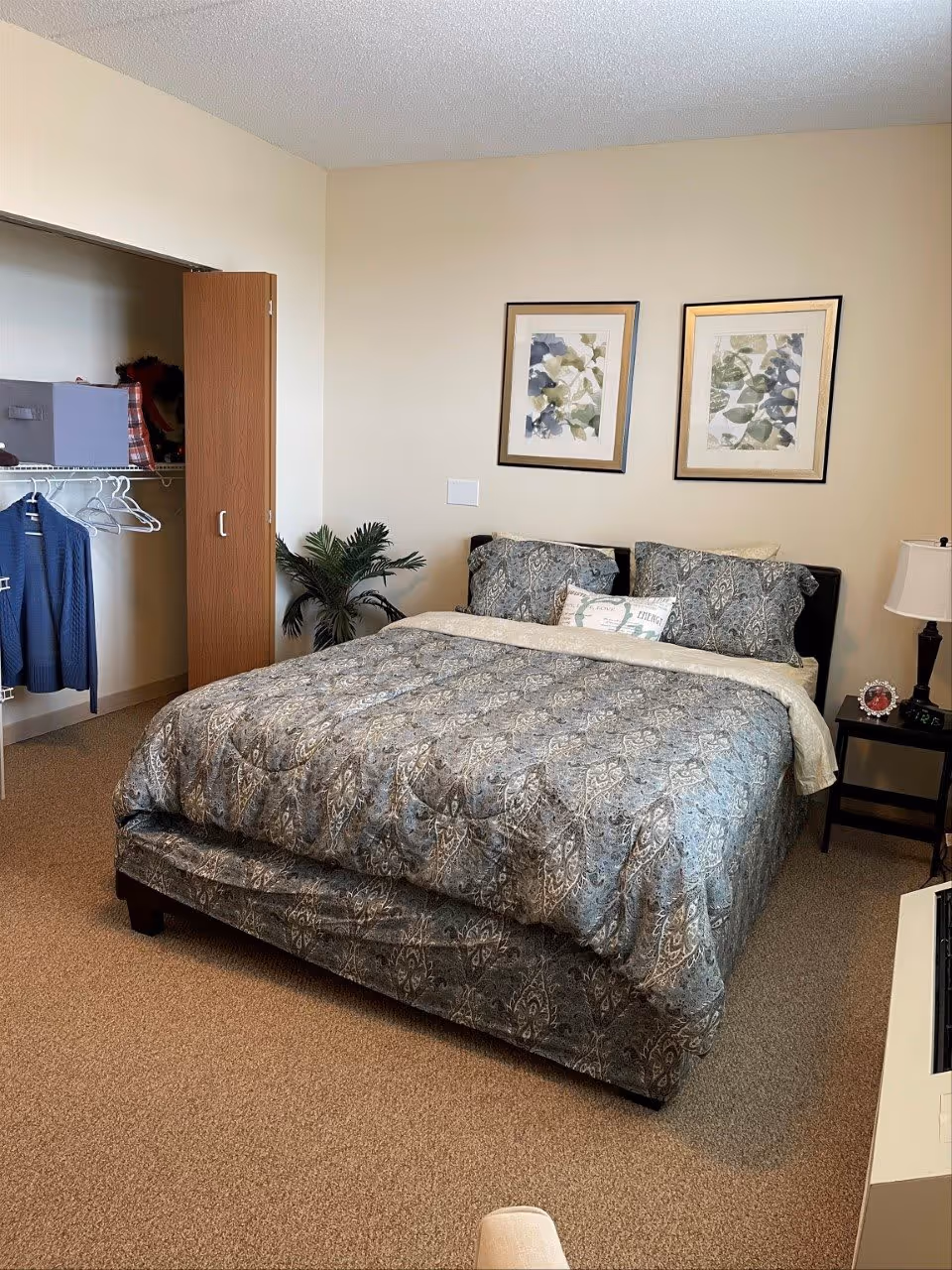 A bedroom with a neatly made bed featuring a patterned gray and beige comforter and matching pillows. Above the bed are two framed botanical prints. To the left is an open closet with hangers and a blue jacket hanging inside. A small green plant is placed near the closet. To the right of the bed is a nightstand with a lamp and a small clock. The room has beige walls and carpeted floor.
