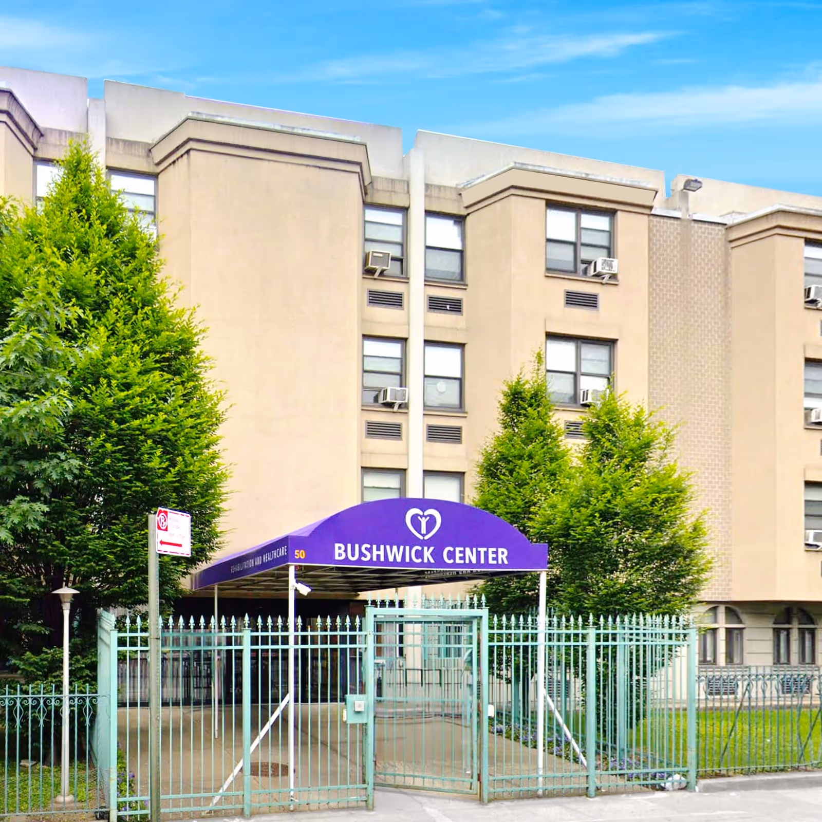 Exterior view of Bushwick Center, a multi-story beige building with several windows and air conditioning units. The entrance is marked by a purple canopy with the name 'Bushwick Center' and a heart logo. There is a green metal fence and gate in front of the entrance, along with two green trees on either side of the canopy.