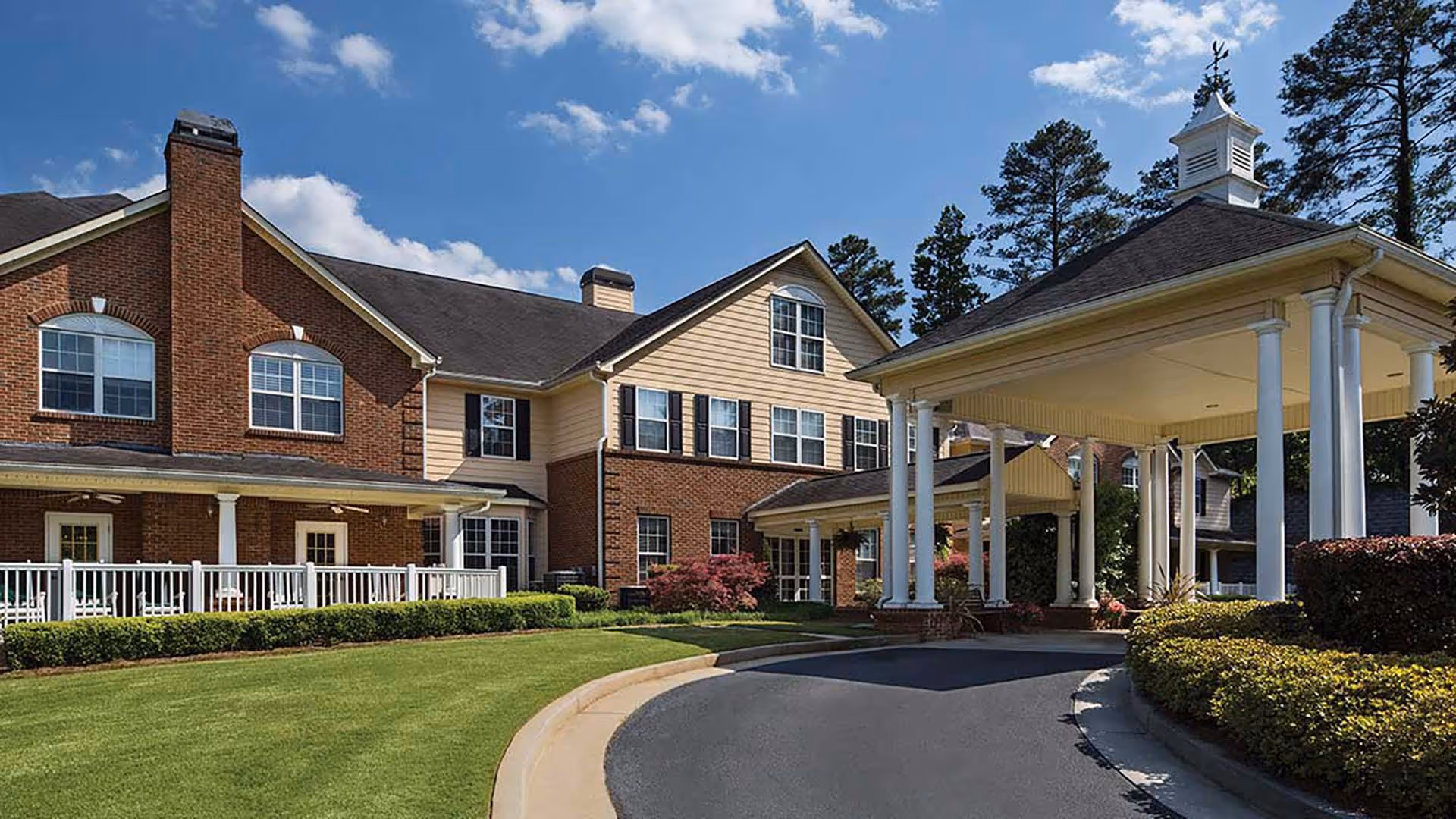 Exterior view of Atria Park of Tucker showing a large two-story building with brick and beige siding, multiple windows, and a covered entrance with white columns. The area is landscaped with green grass, bushes, and trees under a partly cloudy blue sky.