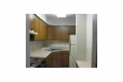 Small kitchen area with wooden cabinets, a white refrigerator, a stove with an oven, and a countertop with a sink. The walls are light-colored and the ceiling has a fluorescent light fixture.