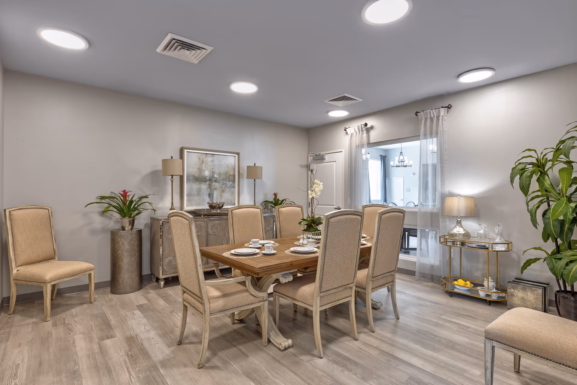 A well-lit dining room with a wooden dining table set for six, surrounded by beige upholstered chairs. The room features light wood flooring, neutral-colored walls, two table lamps on a sideboard, potted plants, a small bar cart with glassware, and a window with sheer curtains looking into another room.