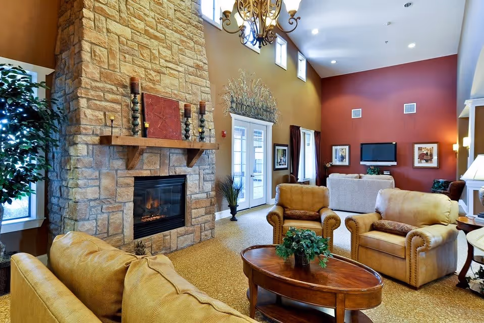 A cozy living room area in an assisted living facility featuring a large stone fireplace with candles and decorative items on the mantel. The room has high ceilings with a chandelier, multiple windows letting in natural light, and comfortable seating including leather armchairs and sofas arranged around a wooden coffee table with a small plant. The walls are painted in warm tones of brown and red, and there is a flat-screen TV mounted on the far wall with framed artwork on either side.