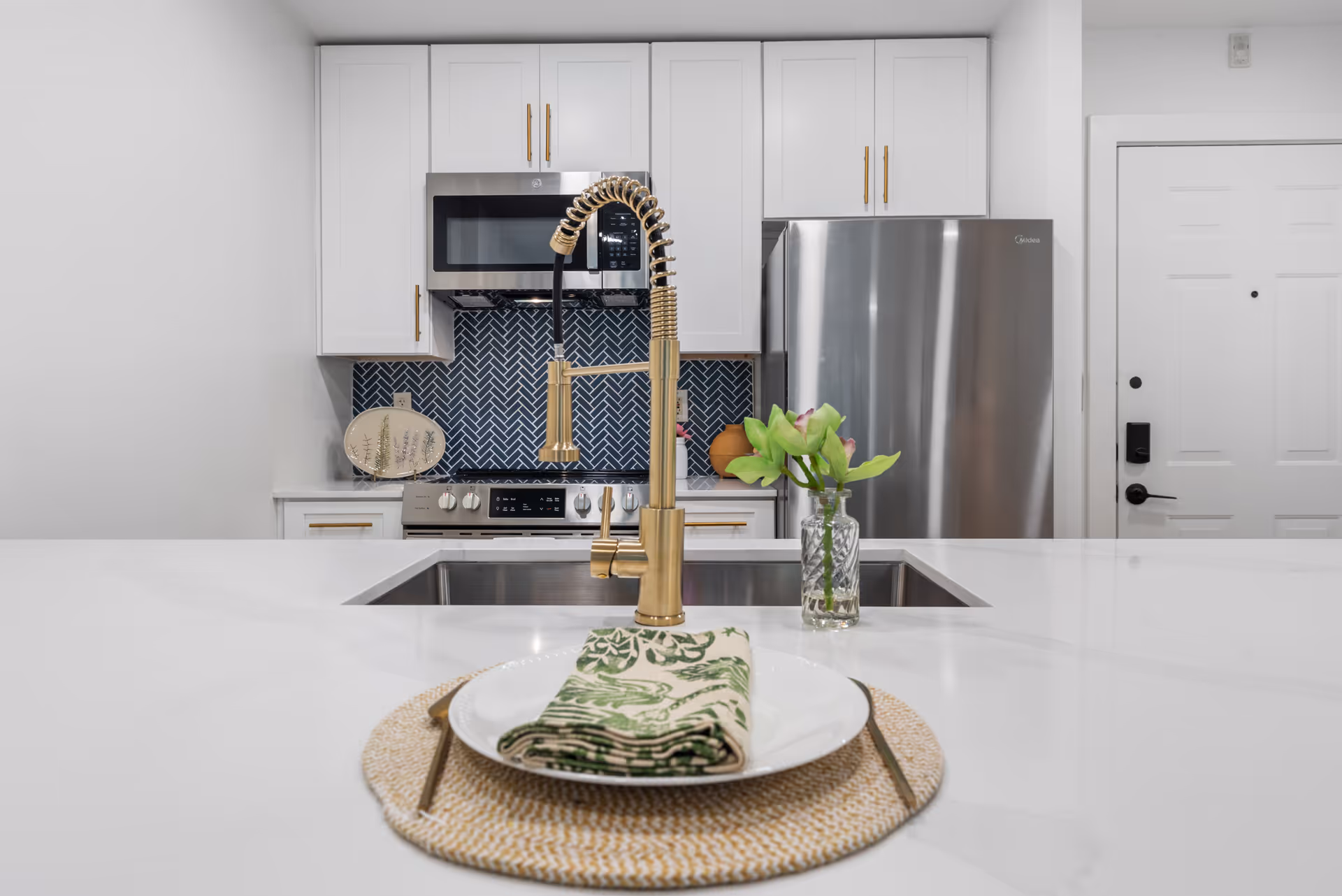 Modern kitchen with white cabinets, a stainless steel refrigerator and microwave, a stove with a blue herringbone tile backsplash, and a gold faucet over a sink in the foreground. A plate with a green patterned napkin and a small vase with green flowers are on the white countertop.