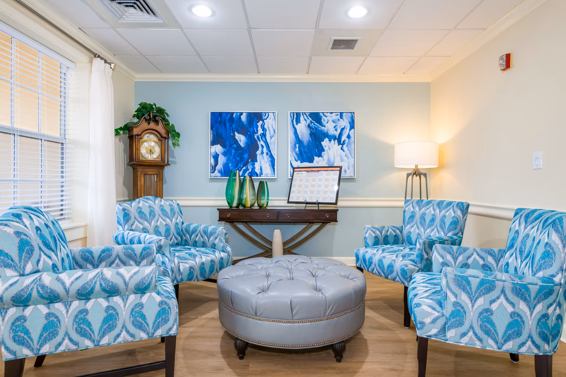 A cozy sitting area with four blue patterned armchairs arranged around a round, tufted gray ottoman. Behind the chairs is a wooden console table with three green glass vases and a framed document. On the wall above the table are two abstract blue and white paintings. To the left is a tall wooden grandfather clock with greenery on top, and to the right is a floor lamp with a white shade. The room has light blue and beige walls, a window with white curtains, and a wooden floor.