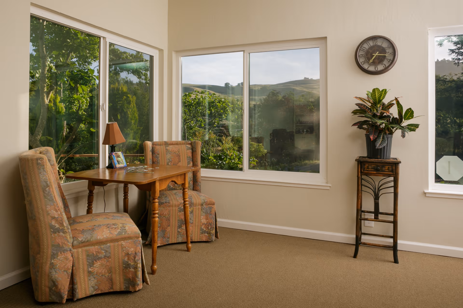 A cozy corner of a living room with two upholstered chairs featuring floral patterns around a small wooden table. On the table is a lamp and a framed photo. Large windows behind the table show a view of green trees and hills outside. A small wooden plant stand with a potted plant is positioned against the wall, and a round wall clock hangs above it.