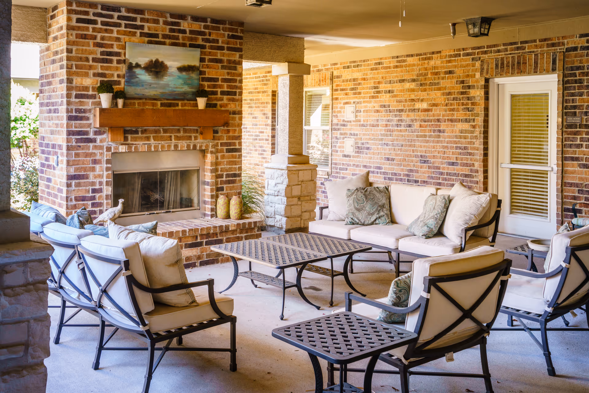 Covered outdoor seating area with cushioned chairs and sofas arranged around two metal tables in front of a brick fireplace. The fireplace has a wooden mantel with small potted plants and a landscape painting above it. The space is enclosed by brick walls and has a door with a window and blinds.