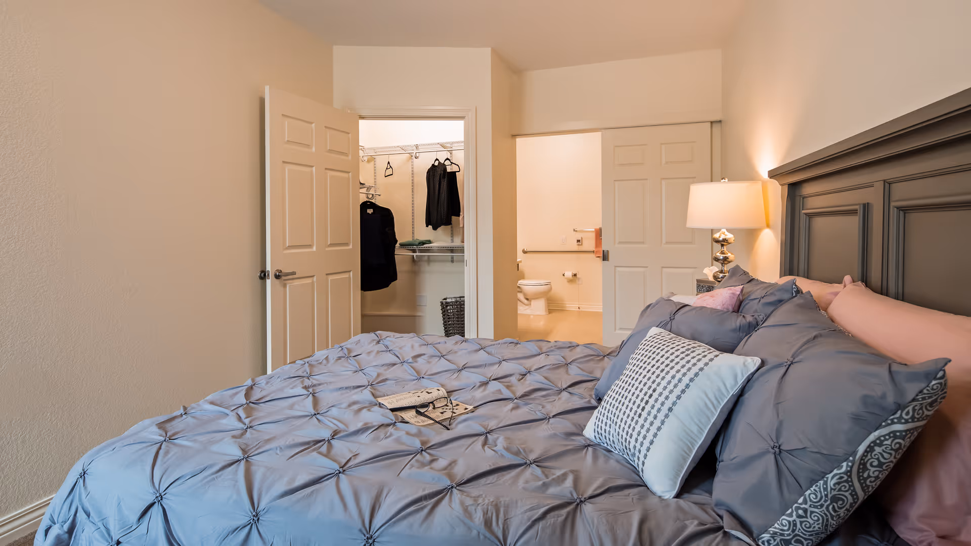 A bedroom with a neatly made bed featuring multiple pillows and a blue comforter. A bedside table with a lamp is next to the bed. The room has an open door leading to a walk-in closet with clothes hanging and another sliding door opening to a bathroom with a visible toilet and grab bars.