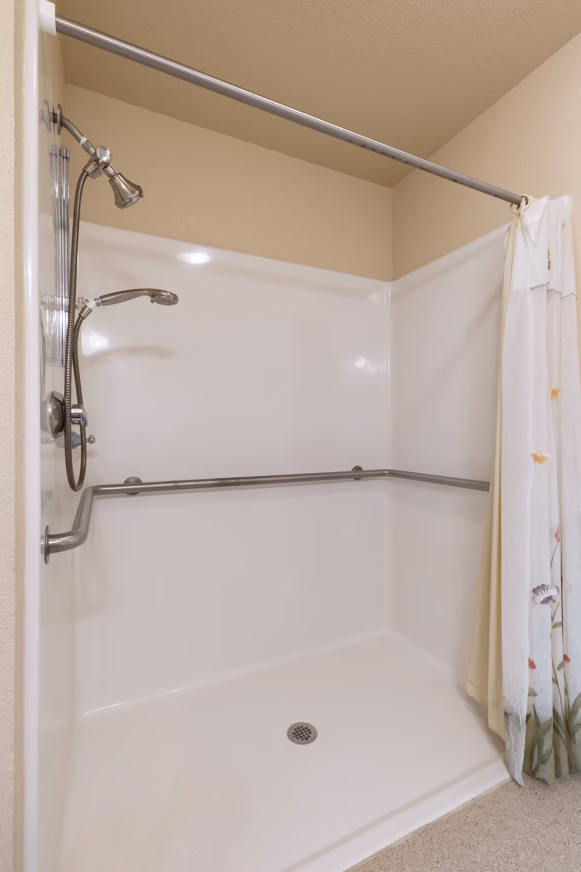 White walk-in shower with stainless steel grab bars, a handheld showerhead and a floral shower curtain.