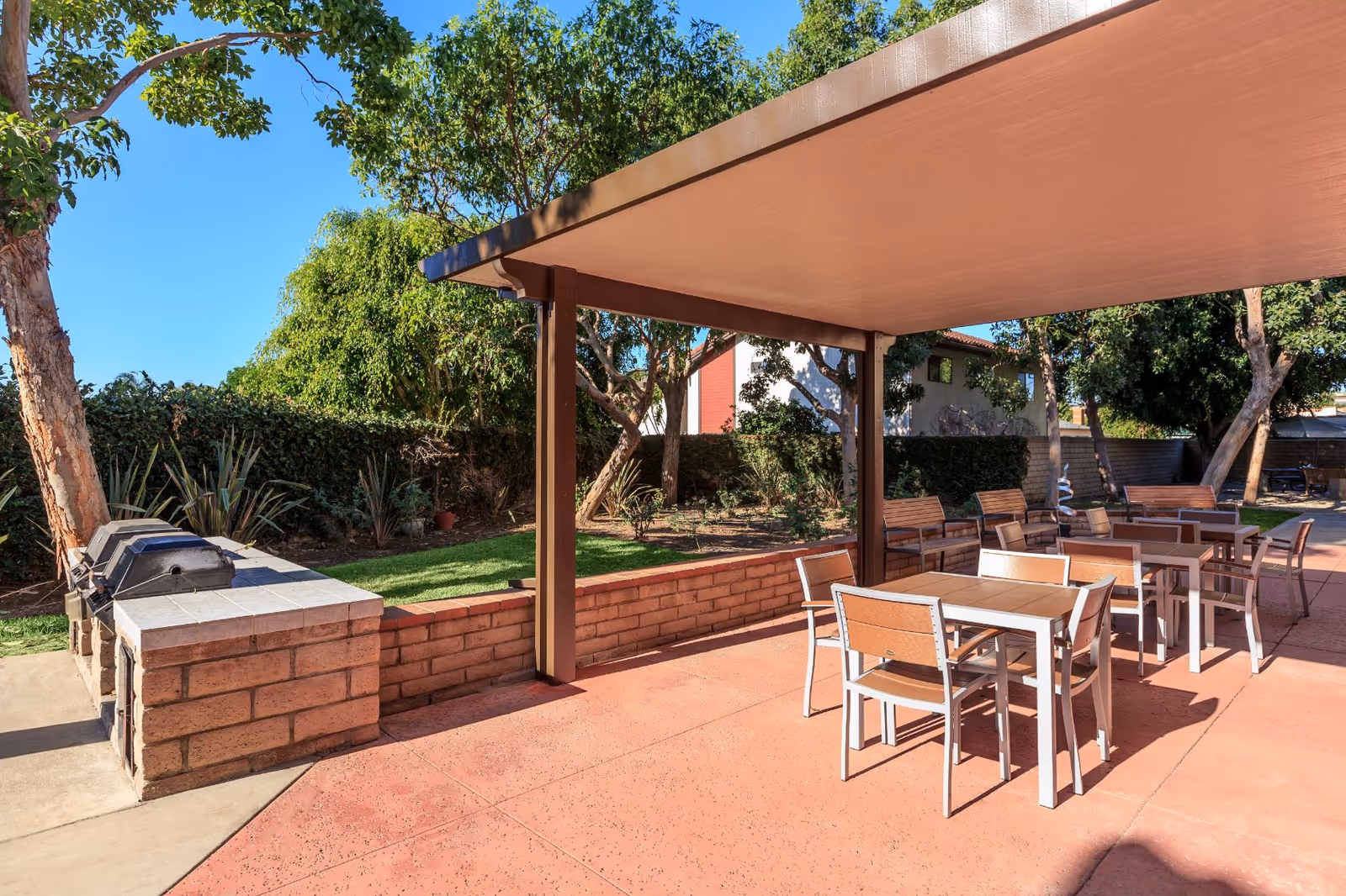 Outdoor patio area with several tables and chairs under a large canopy. There is a brick barbecue grill on the left side and green trees and bushes surrounding the space under a clear blue sky.
