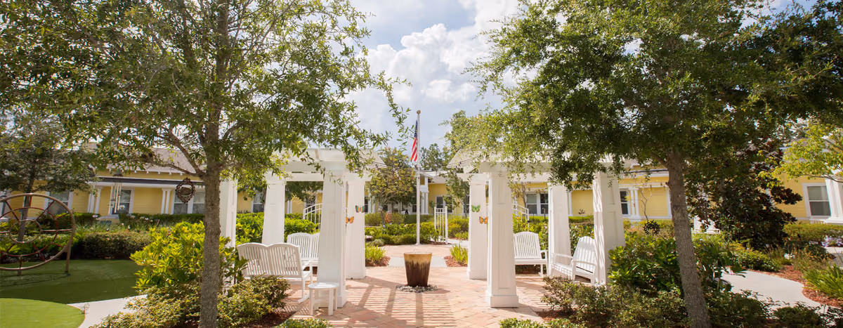Sunny landscaped courtyard with a white pergola and benches, trees, and a yellow single-story building in the background.