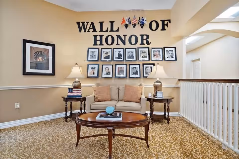 A cozy seating area with a beige loveseat and two matching pillows, flanked by two wooden side tables with lamps. Above the loveseat is a 'Wall of Honor' display featuring framed photographs of veterans and small flags. The room has beige walls, patterned carpet, and a white railing on the right side.