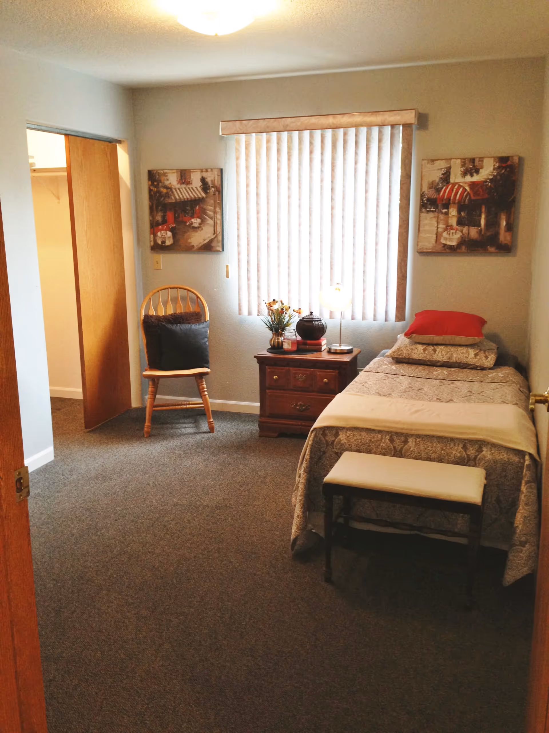 A small bedroom with a single bed covered in patterned bedding and a red pillow. Next to the bed is a wooden nightstand with a lamp, flowers, and decorative items. A wooden chair with a dark cushion is placed near an open closet with sliding wooden doors. The room has carpeted flooring, light-colored walls, vertical blinds on the window, and two framed pictures on either side of the window.