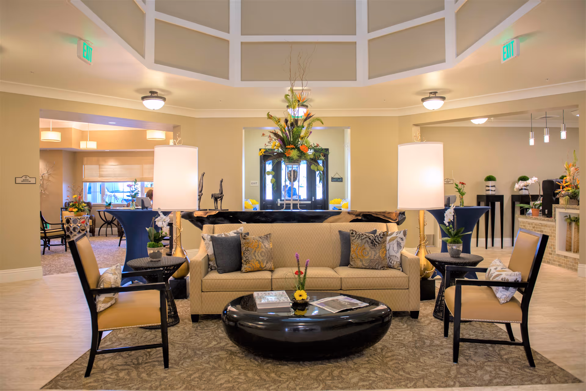 A well-lit, elegant living room area in a senior living facility featuring a beige sofa with decorative pillows, two armchairs with cushions, two tall lamps on side tables, a black oval coffee table with magazines and a small flower arrangement, and a large floral centerpiece on a console table behind the sofa. The room has beige walls, carpeted flooring, and visible adjoining rooms with additional seating and decor.