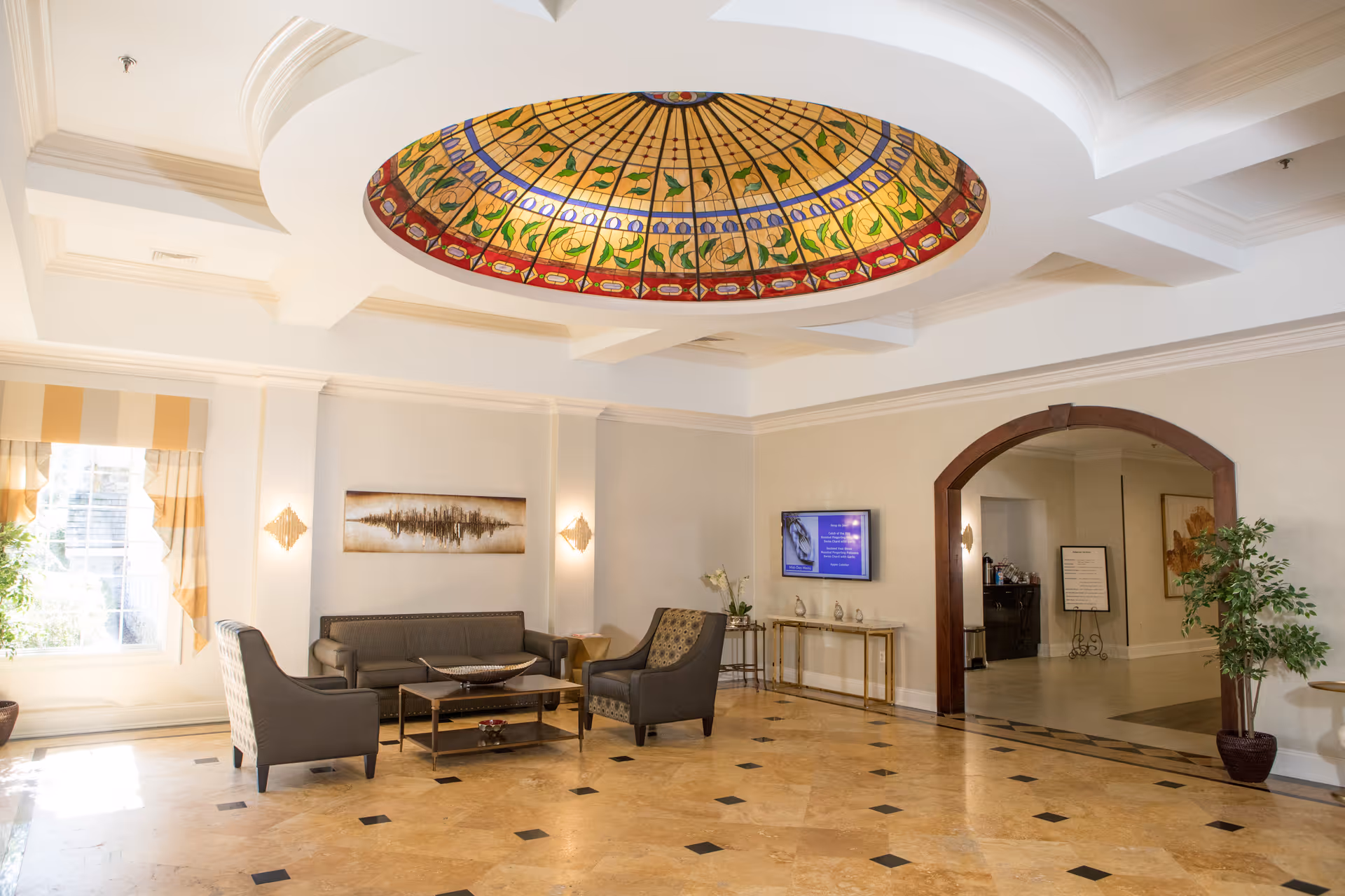 A spacious, well-lit living room area with a stained glass dome ceiling light fixture. The room features a seating arrangement with a sofa and two armchairs around a coffee table. There is a large window with yellow and white striped curtains, a wall-mounted TV, decorative wall sconces, and a potted plant near an arched doorway leading to another room.