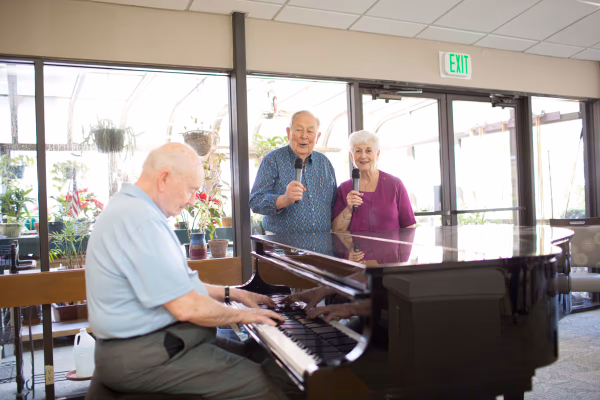 An elderly man playing a grand piano while an elderly man and woman stand beside him holding microphones, inside a bright room with large windows and potted plants.