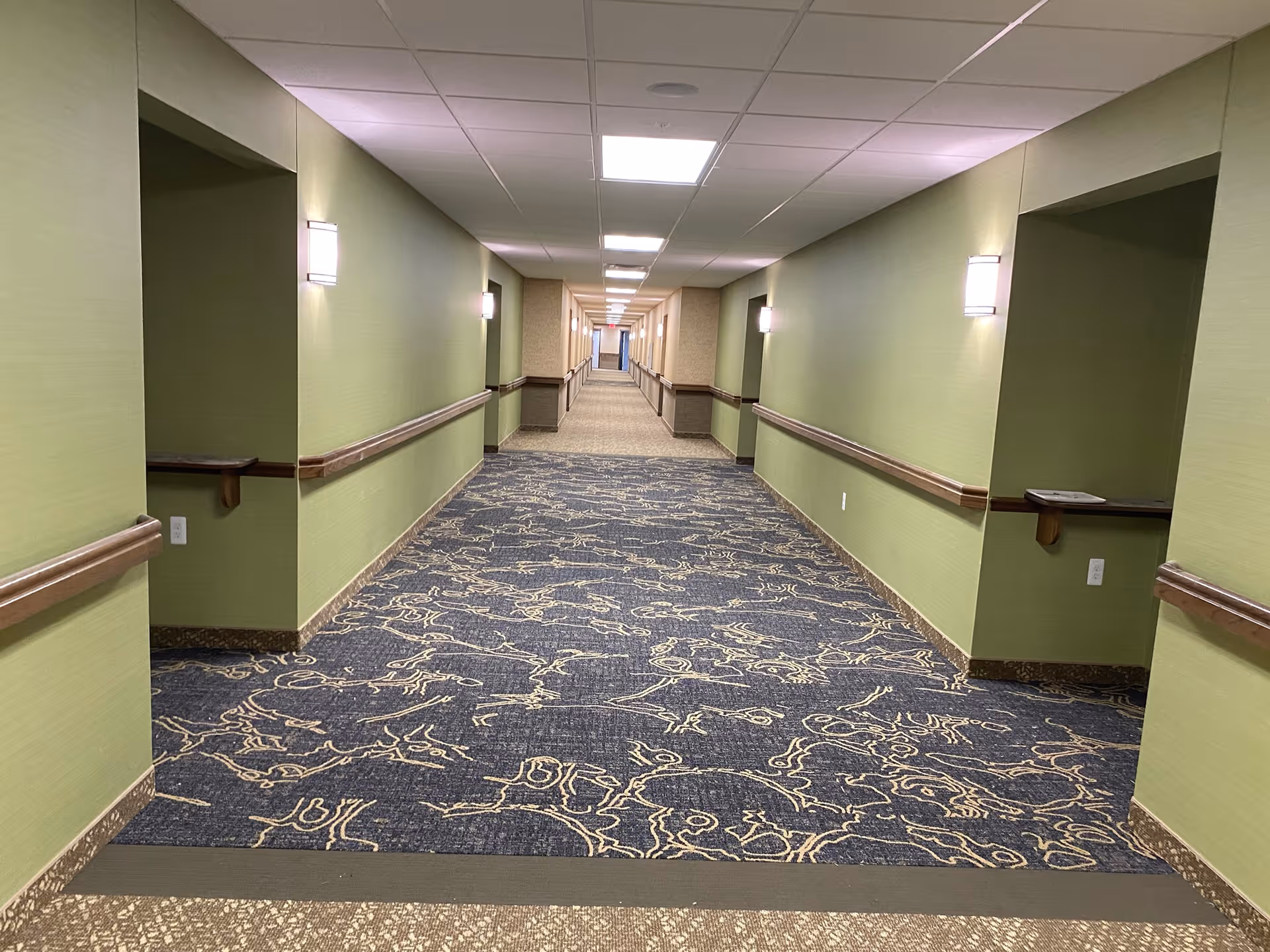 A long, well-lit hallway in a retirement community with green walls, patterned carpet, wooden handrails on both sides, and recessed alcoves with small shelves.