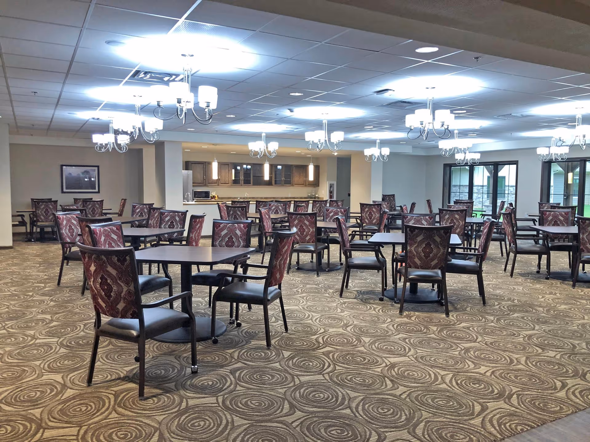A spacious dining room with multiple square tables and patterned chairs arranged neatly. The room features a patterned carpet, several chandeliers hanging from a drop ceiling, and a kitchen area with cabinets and appliances in the background. Large windows on the right side allow natural light to enter.