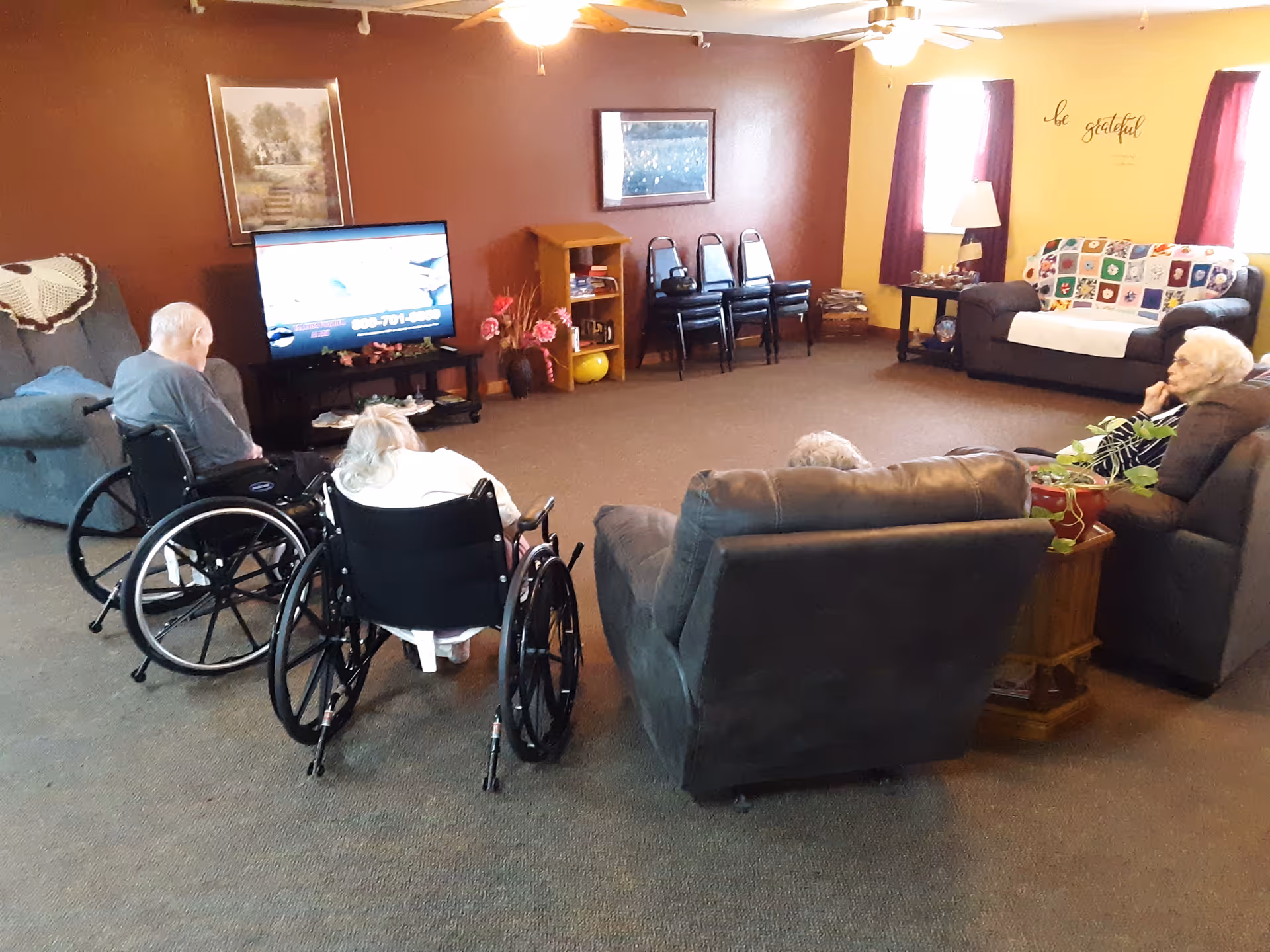 A senior living common room with residents seated in wheelchairs and recliners watching a television.