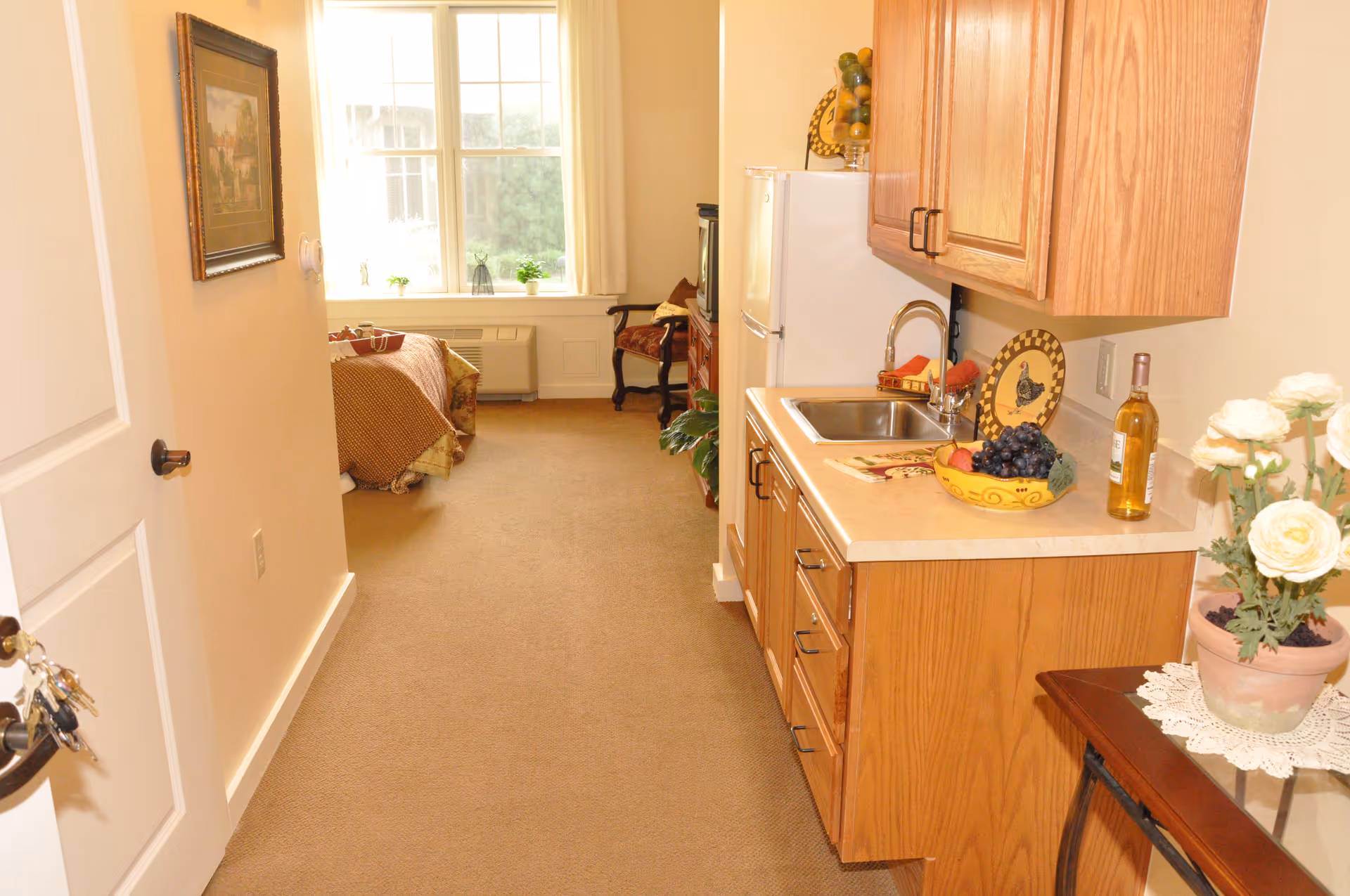 Hallway kitchenette with wooden cabinets and a sink leading to a studio bed by a large window.