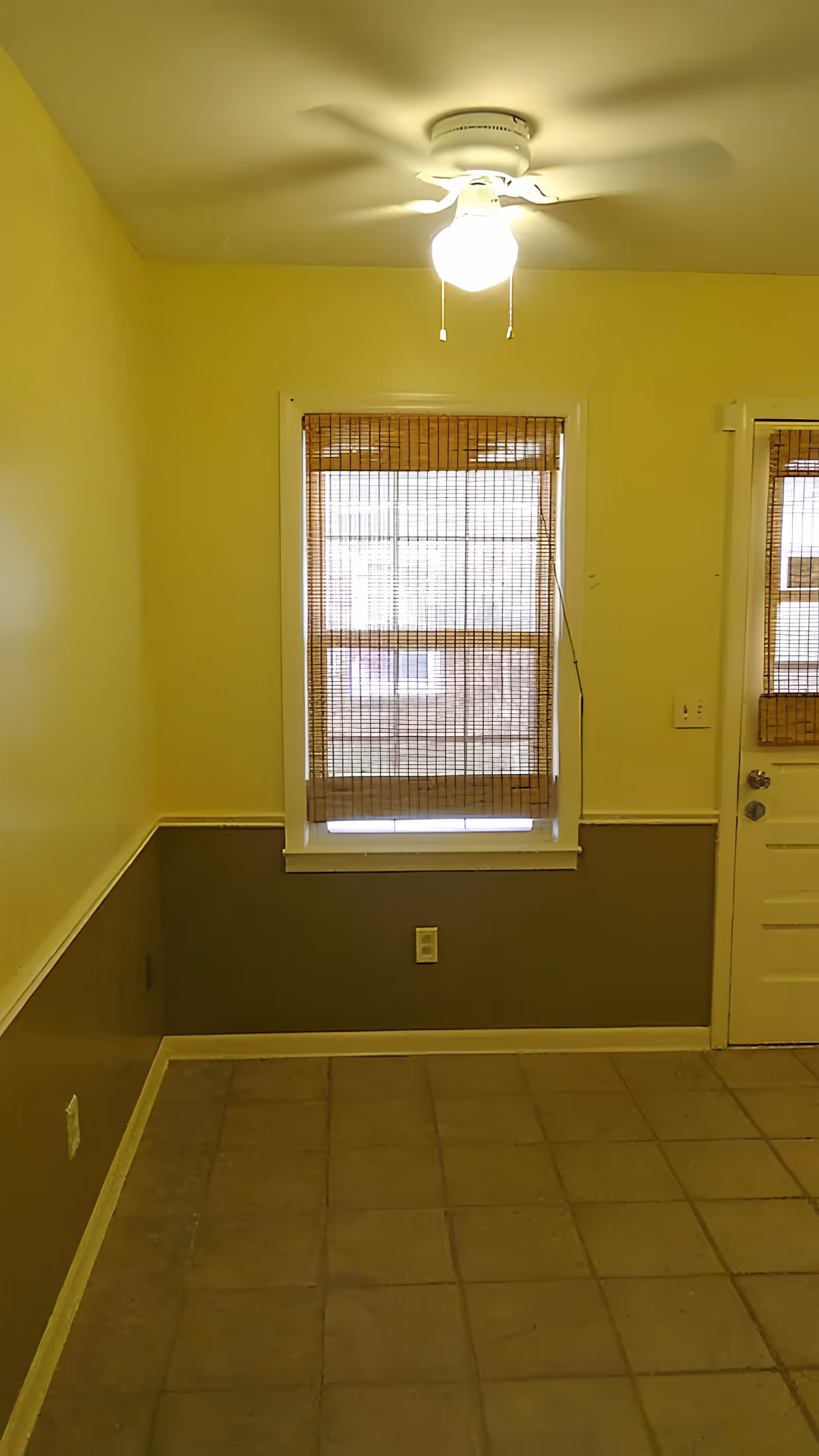 An empty room with yellow walls and a brown lower wall section, a window with bamboo blinds, a white door with a window also covered by bamboo blinds, a ceiling fan with a light turned on, and a tiled floor.