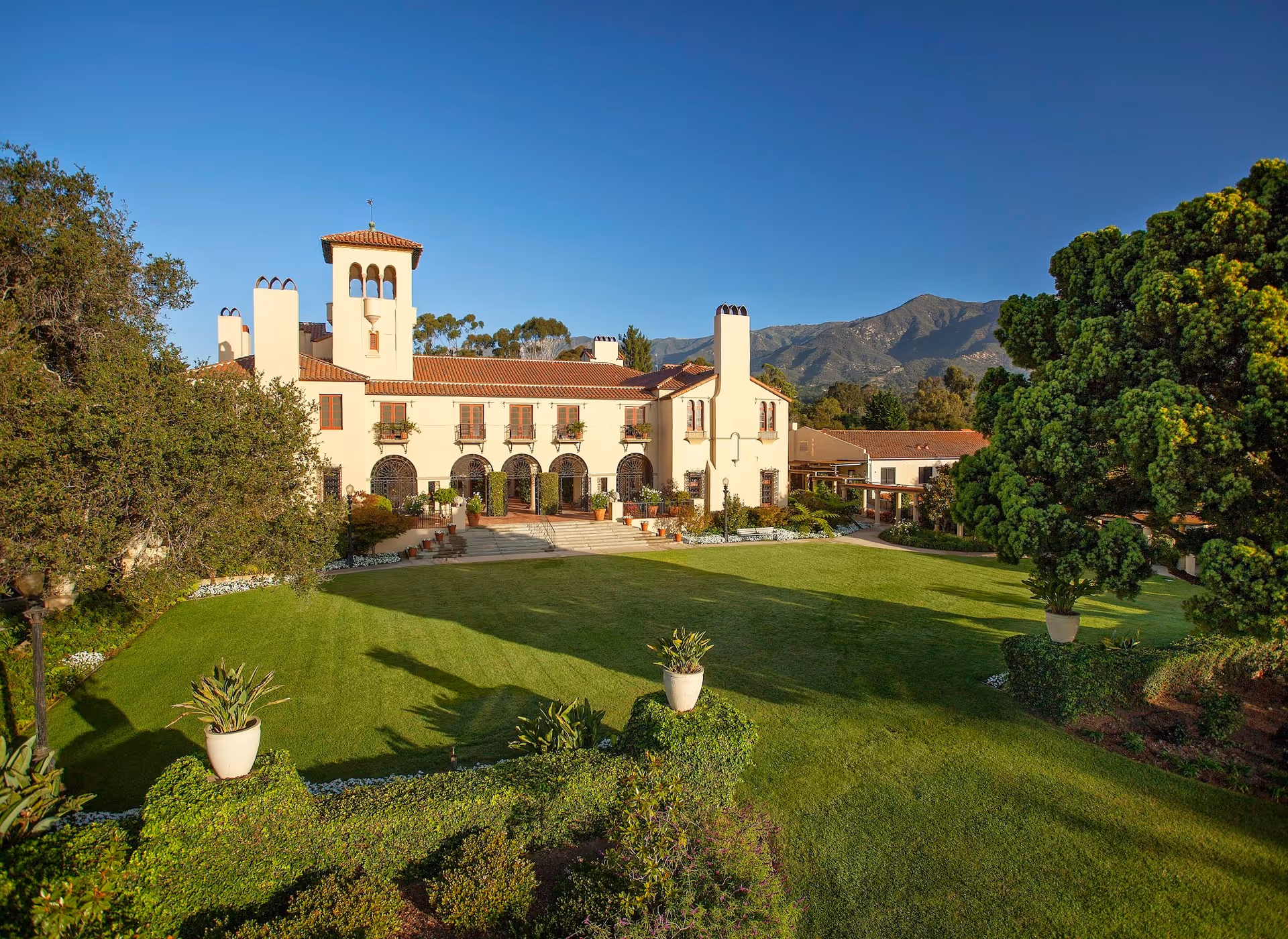A large, elegant building with Spanish-style architecture featuring white walls, red-tiled roofs, and arched windows and doorways. The building is surrounded by a well-maintained green lawn with potted plants and trees. In the background, there are mountains under a clear blue sky.