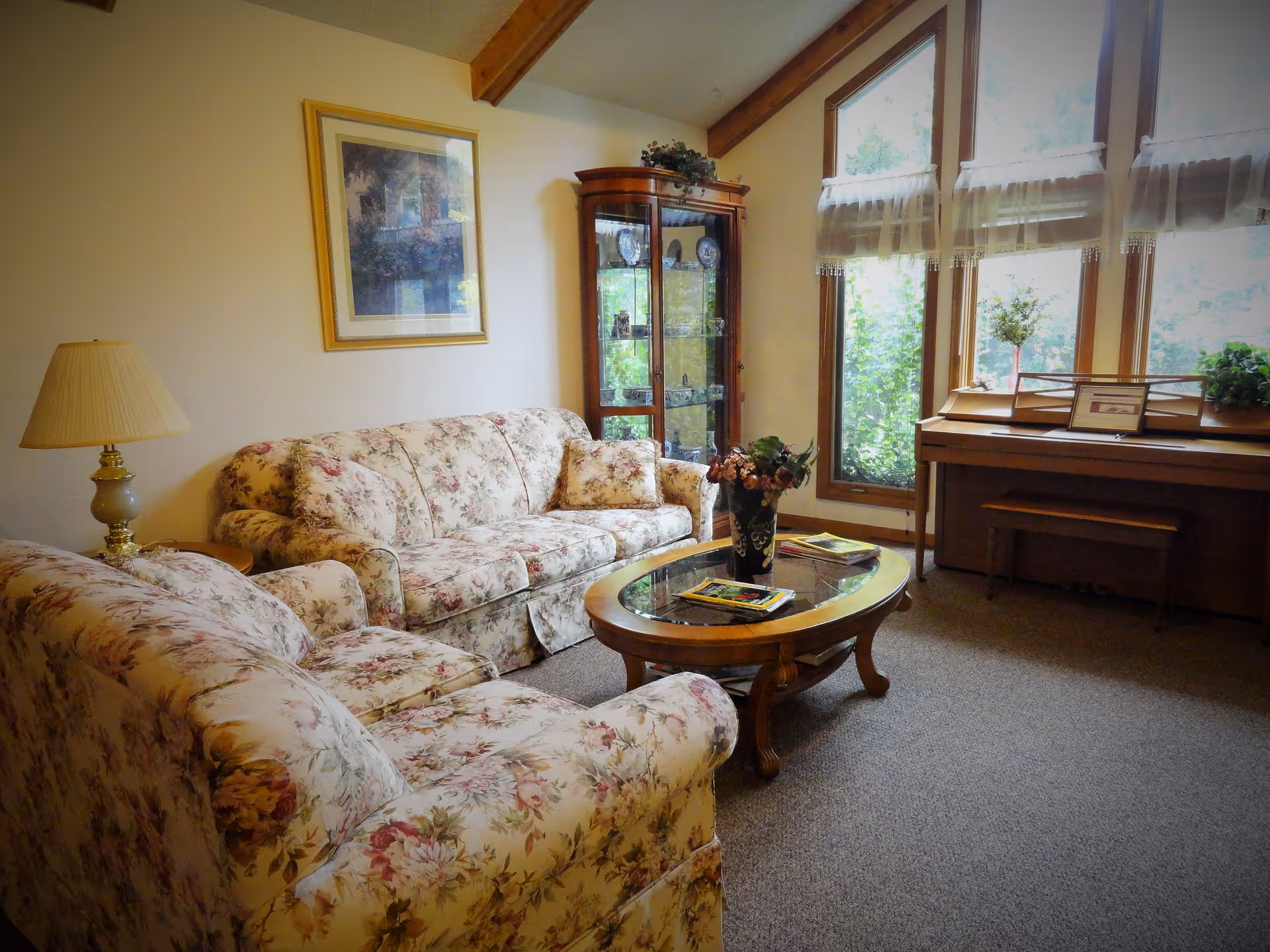 A cozy living room with floral-patterned sofas, a glass-top wooden coffee table with magazines and a flower vase, a wooden display cabinet with decorative items, a piano with a bench, and large windows with sheer curtains letting in natural light.