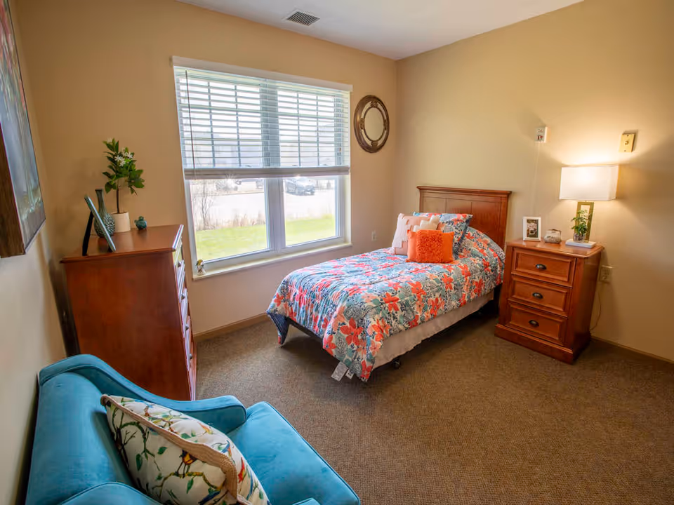 A cozy bedroom with a single bed covered in a floral patterned bedspread with multiple pillows. Next to the bed is a wooden nightstand with a lamp, a small plant, and a framed photo. Across from the bed is a wooden dresser with decorative items and a potted plant. A large window with blinds lets in natural light. A teal armchair with a floral cushion is positioned in the corner of the room. The walls are painted beige and the floor is carpeted.