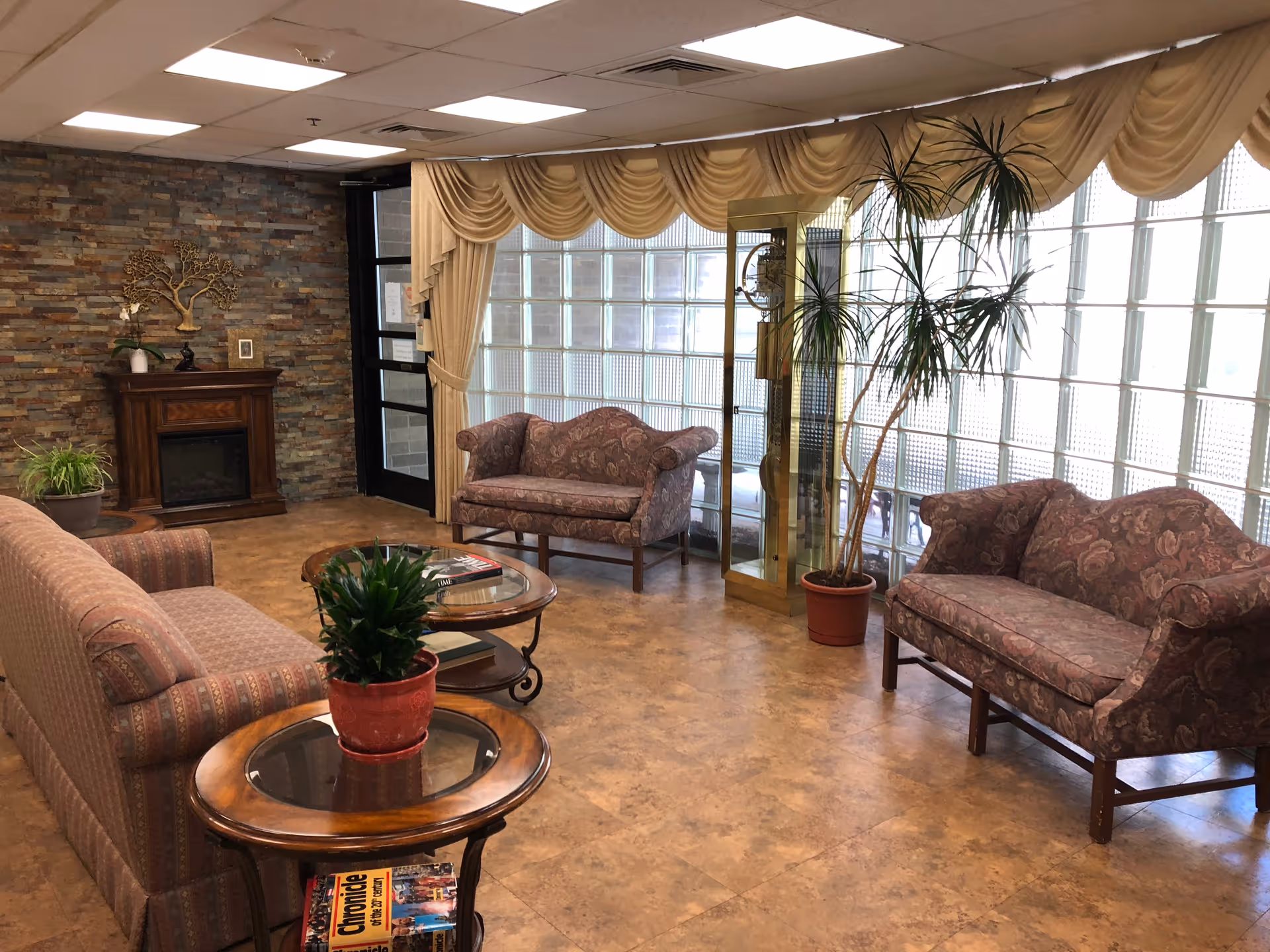 A cozy living room area with patterned upholstered sofas and armchairs arranged around wooden tables with glass tops. There are potted plants on the tables and floor, a decorative fireplace against a stone accent wall, and large glass block windows with beige drapes allowing natural light to fill the space.