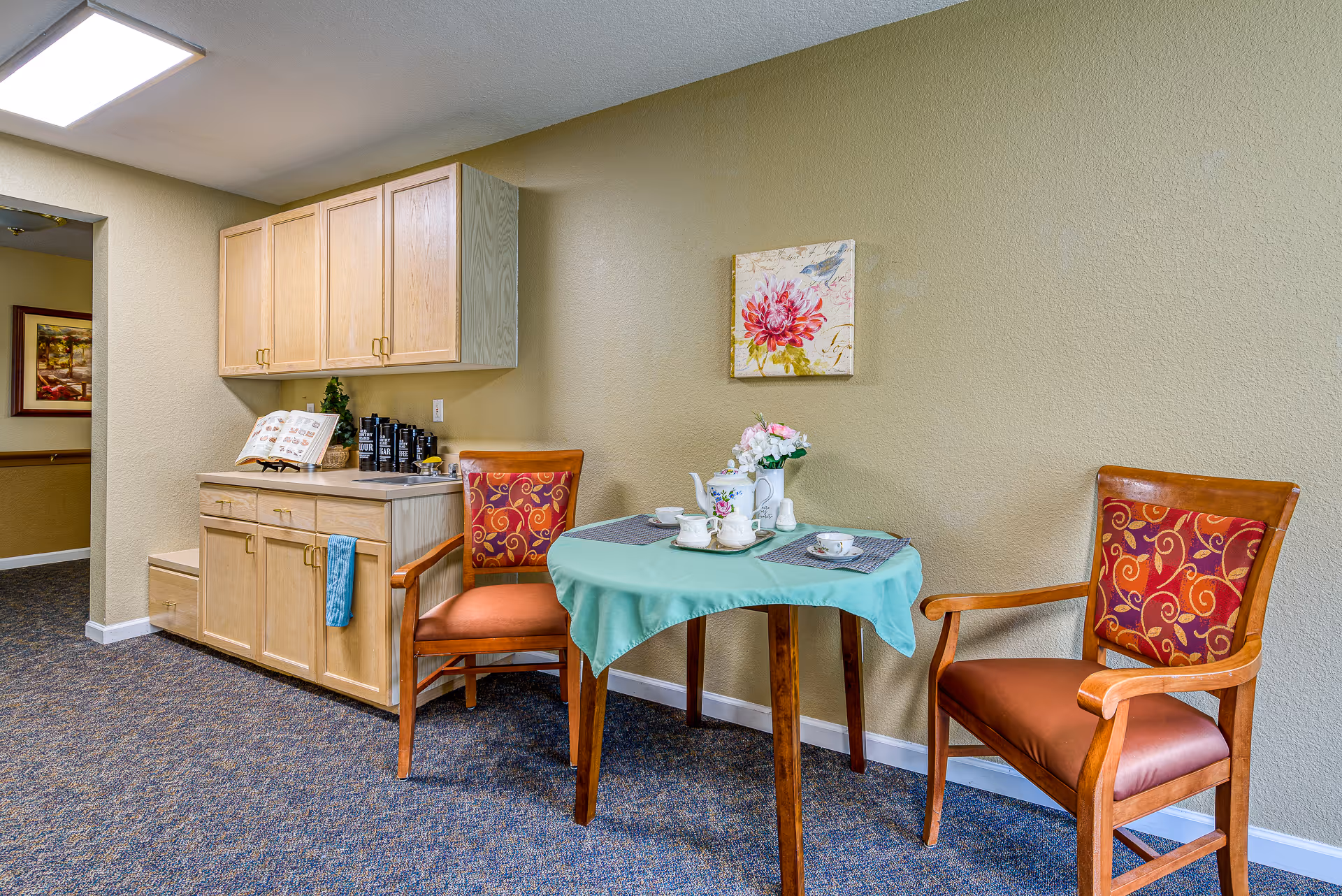 A small dining area with a round table covered with a light blue tablecloth, set with a tea set and a small flower arrangement. Two wooden chairs with orange cushions and patterned backs are placed on either side of the table. Behind the table is a beige wall with a floral painting. To the left, there is a wooden cabinet with upper and lower storage, a countertop with coffee containers, and a blue towel hanging from one of the cabinet handles. The floor is carpeted in a blue patterned design.