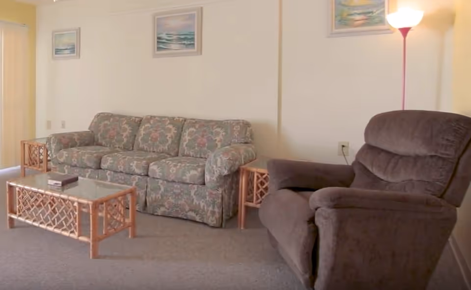 A living room with a floral patterned sofa, a brown recliner chair, a glass-top coffee table, and a side table. There are two framed paintings on the wall and a floor lamp providing light.