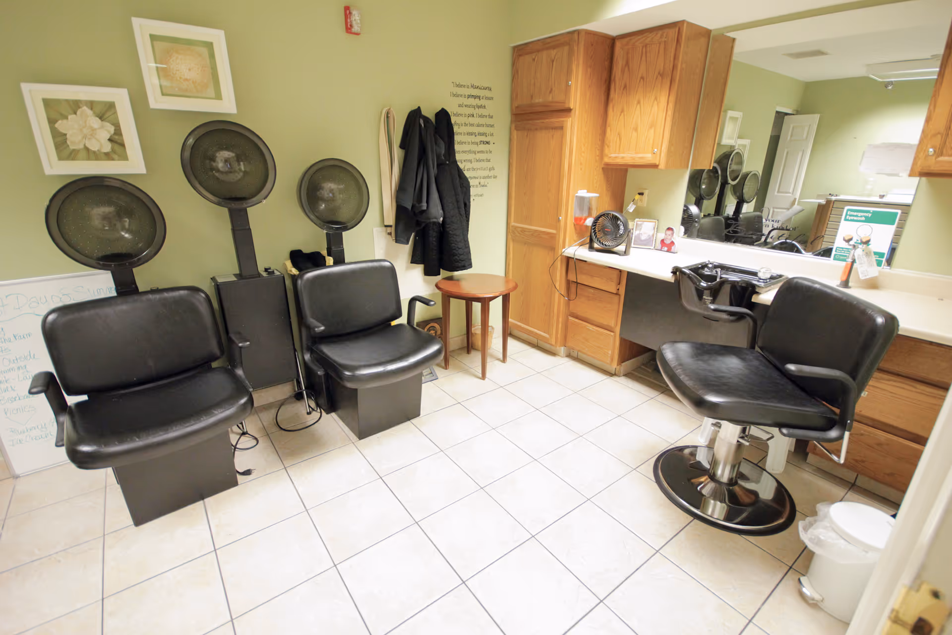 Interior view of a salon area in a senior living facility with three black salon chairs, two hair drying stations, a small wooden table, wooden cabinets, a large mirror, and coats hanging on the wall. The walls are painted light green and the floor is tiled.