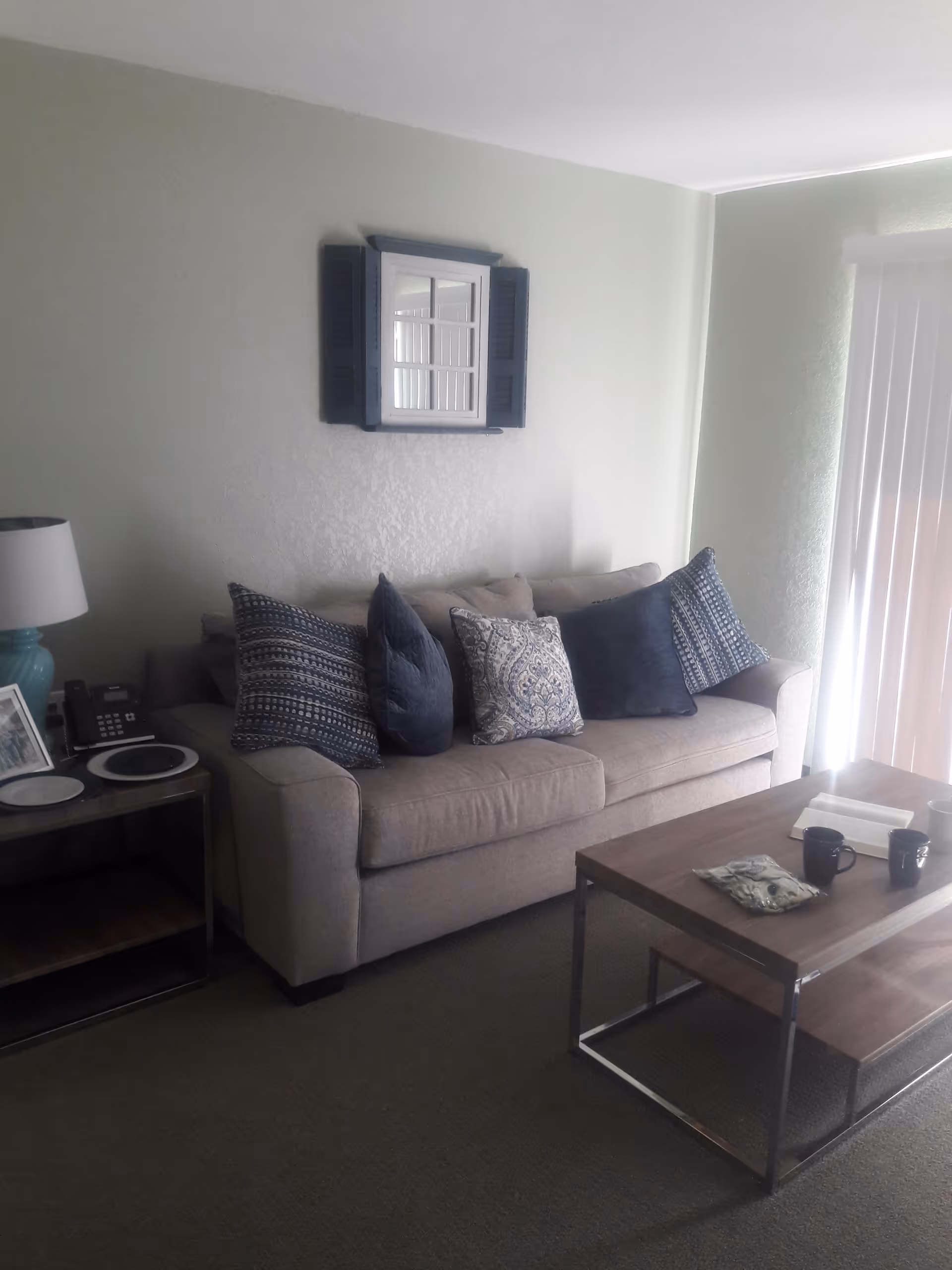A cozy living room with a beige sofa adorned with five decorative pillows in various blue and patterned designs. Above the sofa is a wall-mounted mirror framed with blue shutters. To the left of the sofa is a side table with a blue lamp, a phone, and a framed photo. In front of the sofa is a wooden coffee table with two black mugs, a folded cloth, and a white tray. Vertical blinds cover a window or sliding door on the right side, allowing natural light to enter the room.