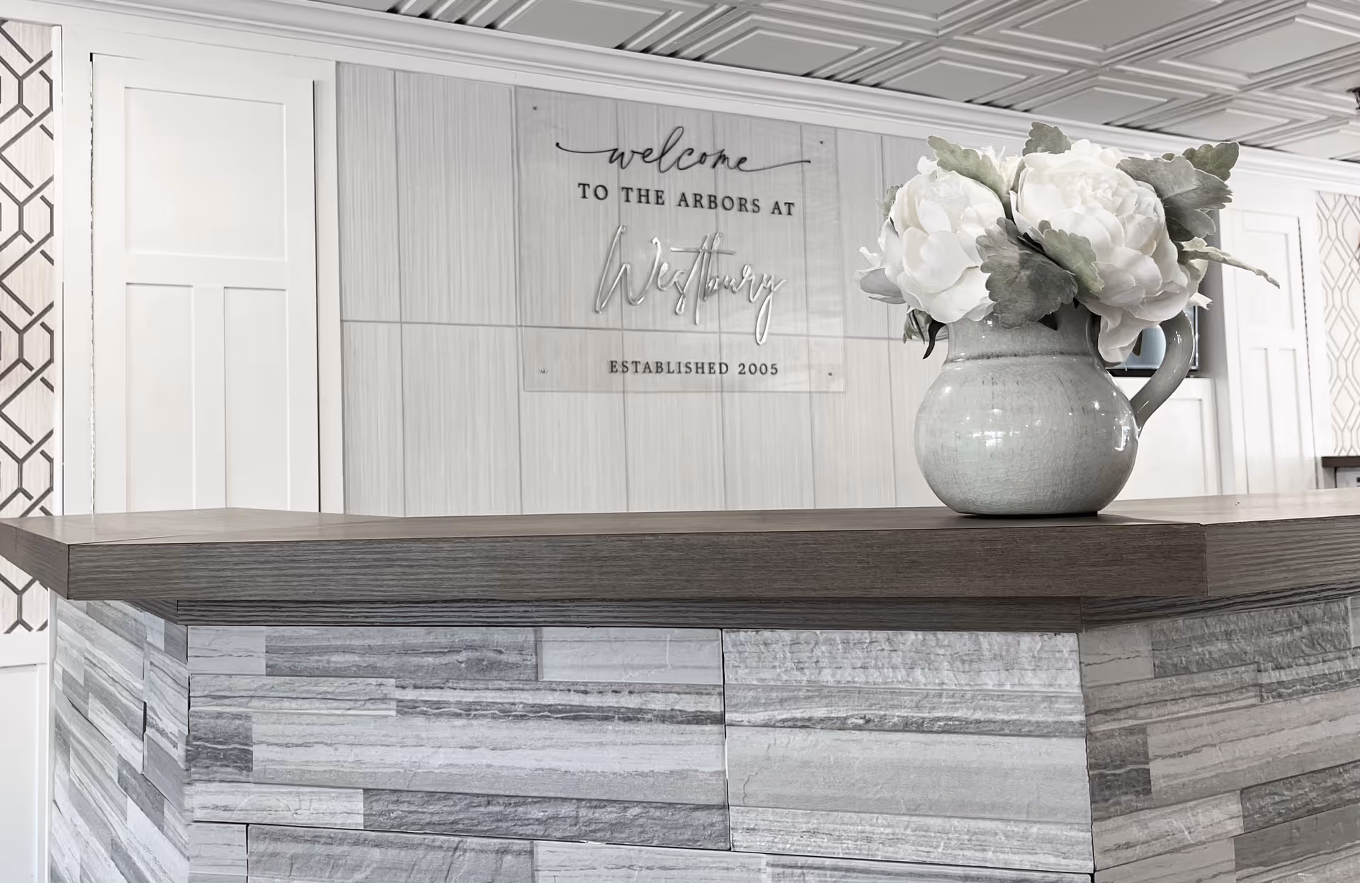 Reception desk and floral arrangement in a lobby with a wall sign that says "Welcome to the Arbors at Westbury".