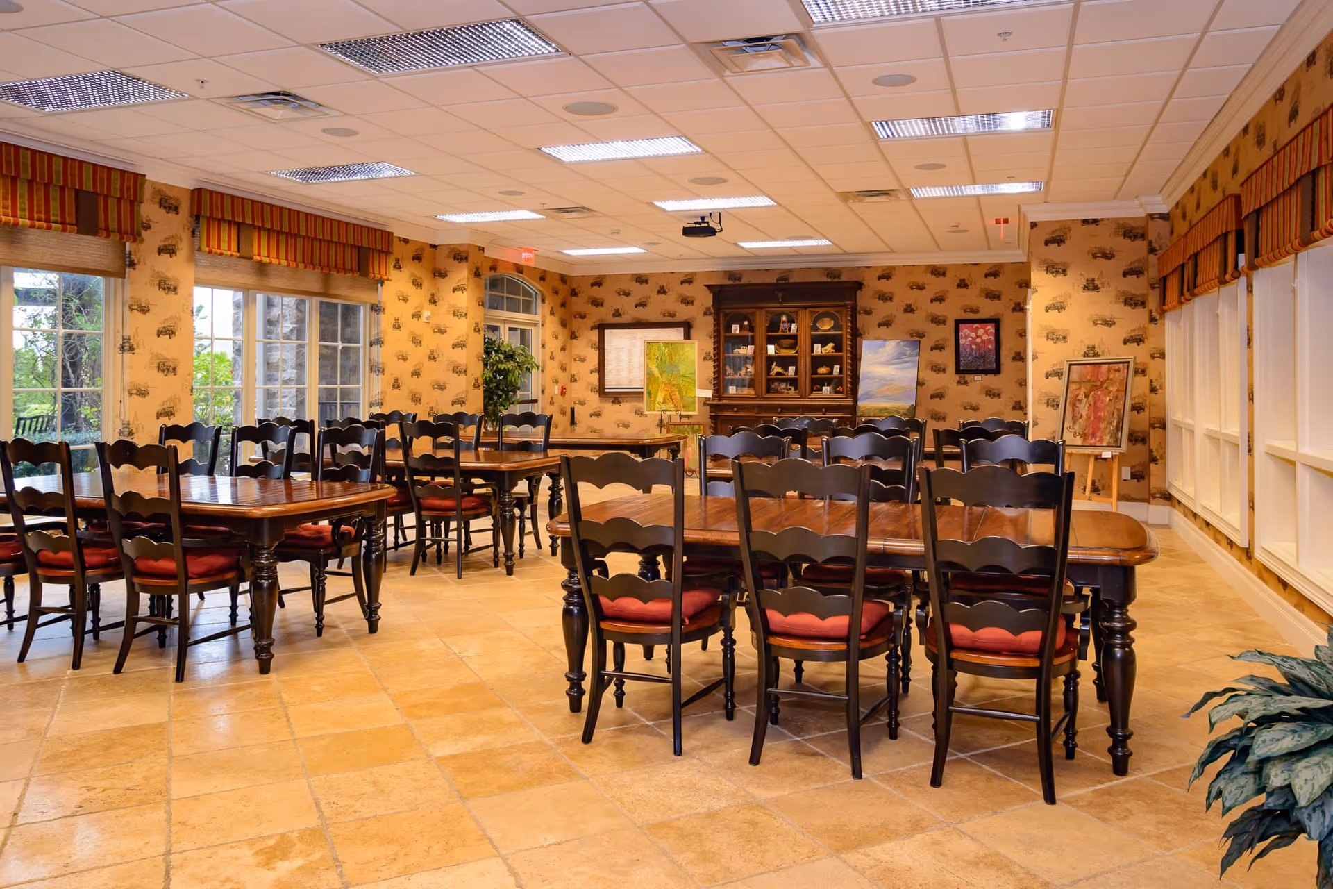 A well-lit dining room with multiple wooden tables and chairs with red cushions. The room has large windows with striped valances, patterned wallpaper, and a wooden cabinet displaying decorative items. There are paintings on easels and framed artwork on the walls.