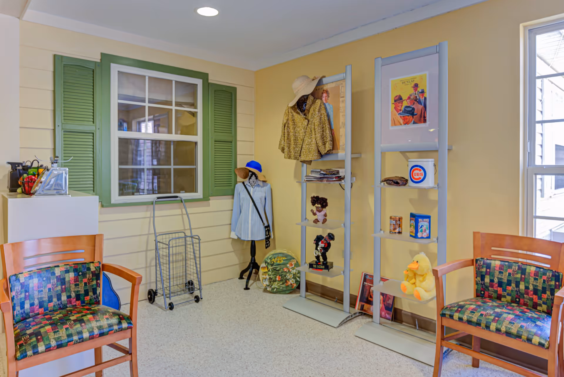 Interior room with two wooden chairs featuring colorful patterned cushions, a window with green shutters, a small white cabinet with decorative items on top, a metal walker, a mannequin dressed in a blue shirt and hat, and two metal display racks holding various decorative objects including framed pictures, dolls, and a stuffed yellow duck. The walls are painted yellow and the floor is light-colored.