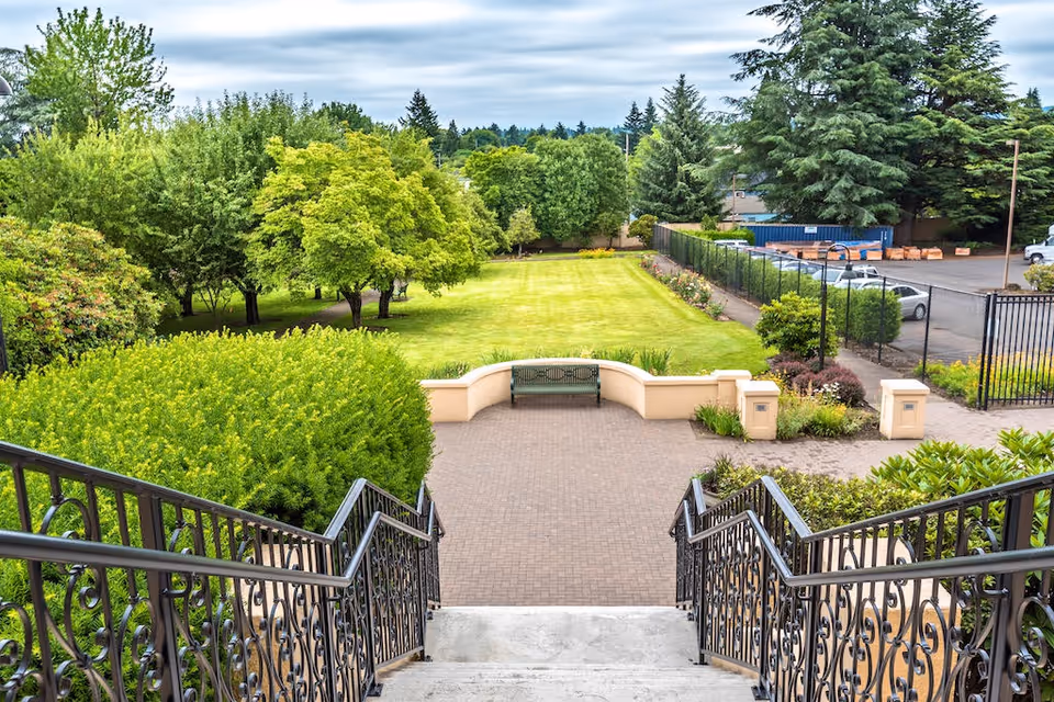 View from the top of a staircase with ornate black railings leading down to a paved patio area with a green bench. Beyond the patio is a well-maintained lawn bordered by various trees and shrubs, with a parking lot and fence visible to the right.