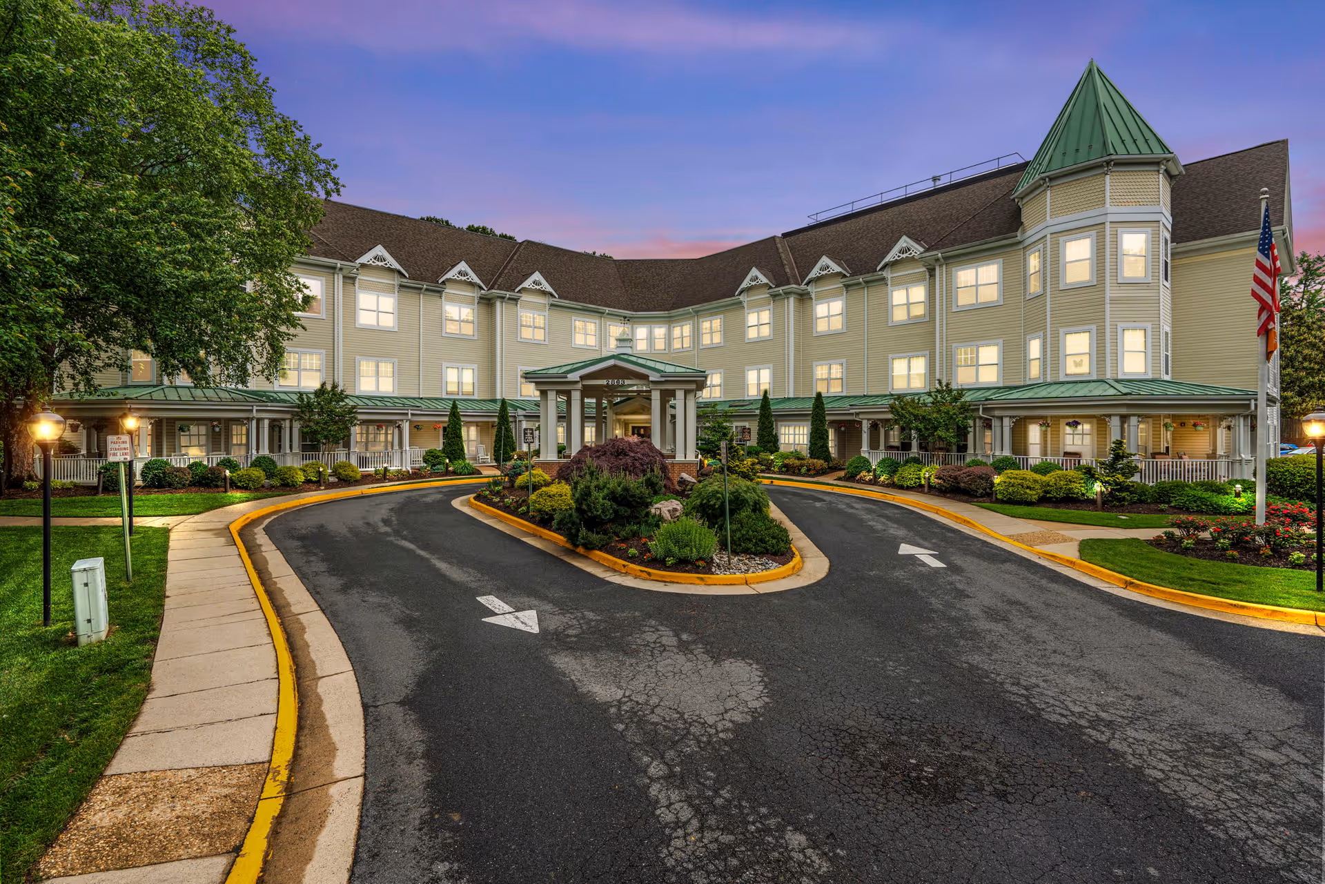 Front entrance of the Sunrise at Hunter Mill senior living building with a circular drive, landscaped center island, and illuminated windows at dusk.