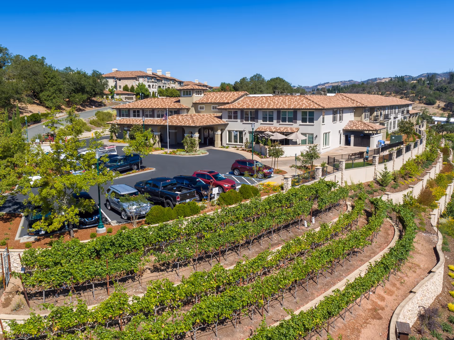A large senior living facility named Villa Capri at Varenna with a terracotta tiled roof, surrounded by landscaped greenery and terraced vineyard rows in the foreground. Several cars are parked in the parking lot near the entrance, and the building is set against a backdrop of hills and clear blue sky.